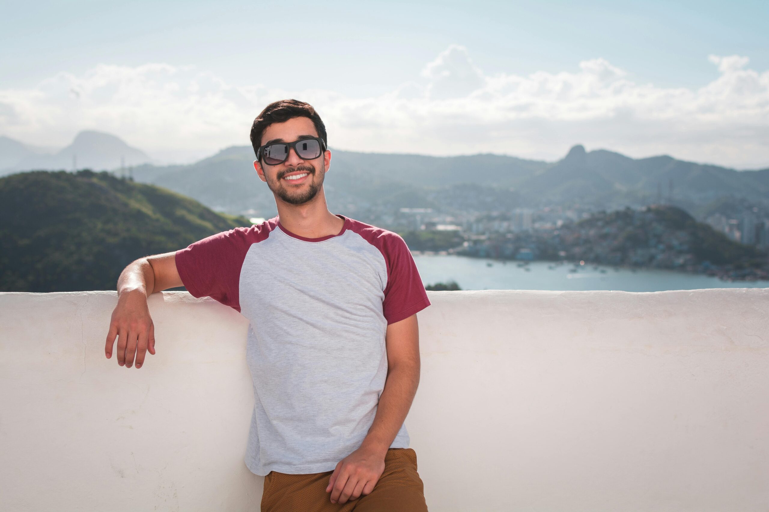 Young man in sunglasses leaning on a balcony with a scenic city view in the background.